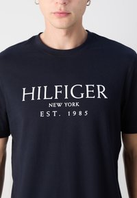 Navy blue cotton t-shirt with short sleeves, featuring white printed text: "HILFIGER NEW YORK EST. 1985" in a bold, uppercase serif font.