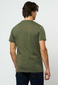 Vyras su tamsiais plaukais, dėvintis alyvuogių žalios spalvos trumpomis rankovėmis marškinėlį ir tamsiai mėlynas džinsines kelnes, stovintis nugara į vienspalvį šviesų foną.