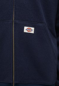 Felpa con zip di colore blu navy realizzata in tessuto testurizzato, con zip dorata e un'etichetta quadrata ricamata nell'area della tasca sinistra.