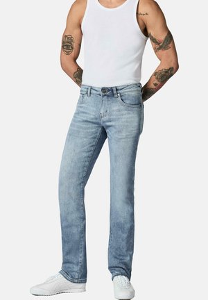 Mann trägt hellblaue Jeans, weißes Tanktop und weiße Sneakers, mit sichtbaren Arm-Tattoos vor weißem Hintergrund.