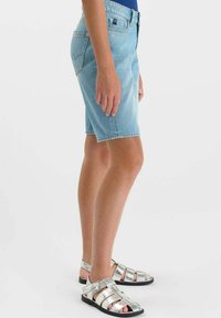 Personne portant un short en jean bleu clair et des sandales argentées à brides métalliques, debout de côté devant un fond uni.