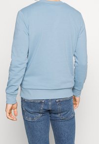 Hombre vistiendo un suéter de manga larga azul claro y jeans de mezclilla azules, mostrado desde la espalda contra un fondo liso.