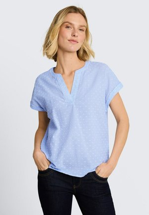 Femme aux cheveux blonds courts portant un chemisier bleu clair texturé à manches courtes et un jean foncé, debout les mains dans les poches devant un fond uni.