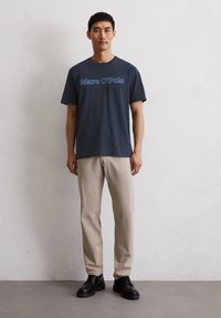 Navyblaue T-Shirt mit dem Aufdruck "Marc O'Polo" in Hellblau, kombiniert mit beigen Hosen und schwarzen Schuhen. Der Stoff wirkt glatt und lässig.