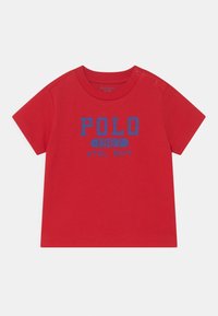 Červené bavlněné tričko s krátkým rukávem, s modrým textem "POLO 1967 ATHL. DEPT." Límec má patentky pro snadné oblečení.