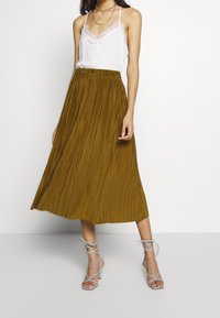 Fållad midikjol i senapsgul, tillverkad av lättviktsmaterial. Matchad med en vit camisole med spetskant och silverfärgade klackar med ankelrem.