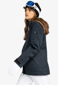 Blauwe waterdichte jas met een rits- en knoopsluiting, verstelbare manchetten en een logopatch op de mouw. Witte wanten en skibril gedragen.