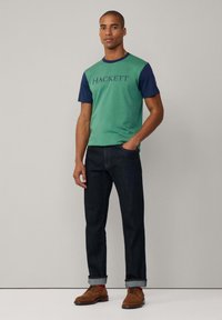 Camiseta de manga corta verde y azul marino con el logo "HACKETT", combinada con jeans de denim oscuro y zapatos de ante marrón, de pie sobre un fondo neutro.