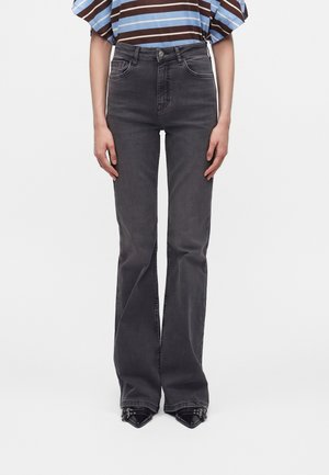 Person iført højtaljede, falmede sorte vide jeans kombineret med sorte spidse sko og en blå og brunstribet skjorte.