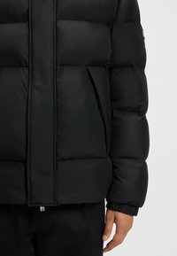 Svart puffjacka med kviltad design, dragkedjor på sidfickorna och elastiska muddar. Tillverkad av ett slätt, väderbeständigt material.