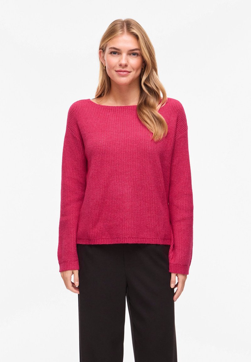 Pull à manches longues et col rond de couleur rose vif, présentant un motif en tricot texturé et une coupe décontractée, associé à un pantalon noir.
