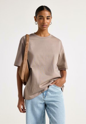 Ung kvinde med øreringe i hoop-stil iført løs beige t-shirt, lyseblå jeans og tan skuldertaske mod ensfarvet baggrund.