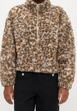 Brązowy i beżowy wzorzysty, miękki sweter z półzamkiem i złotym uchwytem na suwaku, noszony z wysokim stanem czarnych spodni.