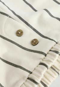 Tessuto a righe crema con strisce orizzontali grigie, due bottoni marroni impreziositi con la scritta "NEXT" e una finitura arricciata.