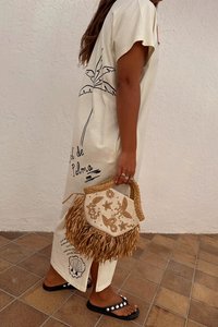Abito largo di colore crema con grafica di palma nera, abbinato a una borsa beige e di paglia. Sandali neri con dettagli bianchi.