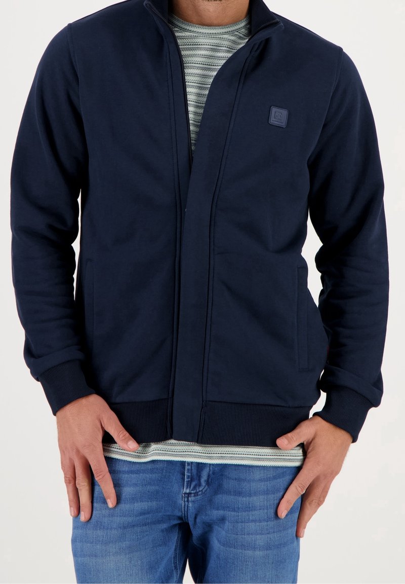Homme portant une veste zippée bleu marine foncé sur une chemise rayée et un jean bleu, debout les mains près des poches devant un fond uni.