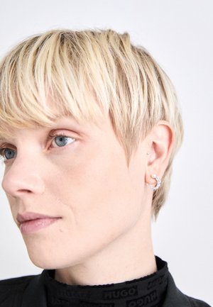 Silverring med en strukturerad design och en pärlaccent, bärs i vänster öra, tillsammans med en kortklippt frisyr.