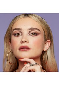 Gros plan sur une personne avec de longs cheveux blonds lisses, présentant un maquillage audacieux, de grandes boucles d'oreilles en or, et des ongles soigneusement manucurés avec des pointes orange.