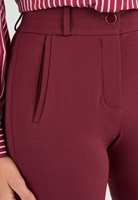 Pantalones tailored burdeos con una tela texturizada, pliegues delanteros y un cierre de botón pulido en la cintura.
