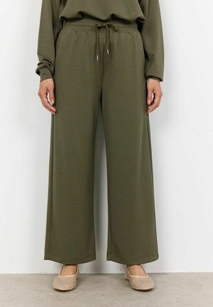 Personne portant un pantalon ample vert olive avec taille à cordon et des chaussures beiges sans lacets, debout contre un mur blanc uni.