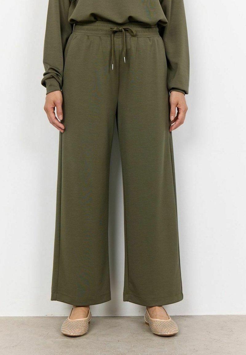 Personne portant un pantalon ample vert olive avec taille à cordon et des chaussures beiges sans lacets, debout contre un mur blanc uni.