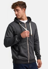 Hoodie zippé en charbon avec une finition texturée, cordons crème, poches avant et poignets côtelés. Design simple avec une coupe décontractée.