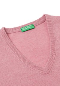Světle růžový svetr s V výstřihem vyrobený z měkké tkaniny. Obsahuje žebrovaný výstřih a švy na ramenou s zelenou etiketou s nápisem "United Colors of Benetton."