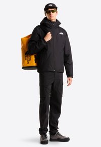 Άνδρας που φοράει μαύρο μπουφάν, παντελόνι, καπέλο και γυαλιά ηλίου, κρατώντας κίτρινο σακίδιο The North Face στον ώμο, στέκεται μπροστά σε λευκό φόντο.