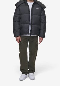 Man wearing a black puffer jacket, white t-shirt, olive cargo pants, and white sneakers standing against a plain background.