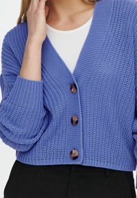 Mujer con un cárdigan de punto azul con grandes botones sobre una blusa blanca, combinado con pantalones negros, con la mano tocando su rostro.