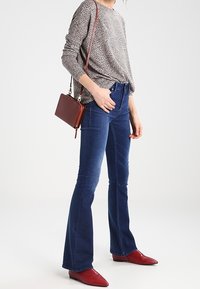 Ausgestellte blaue Jeans in Kombination mit einem langärmeligen Oberteil mit Punkten; eine kleine braune Handtasche tragend. Füße geschmückt mit spitzen roten Stiefeln.