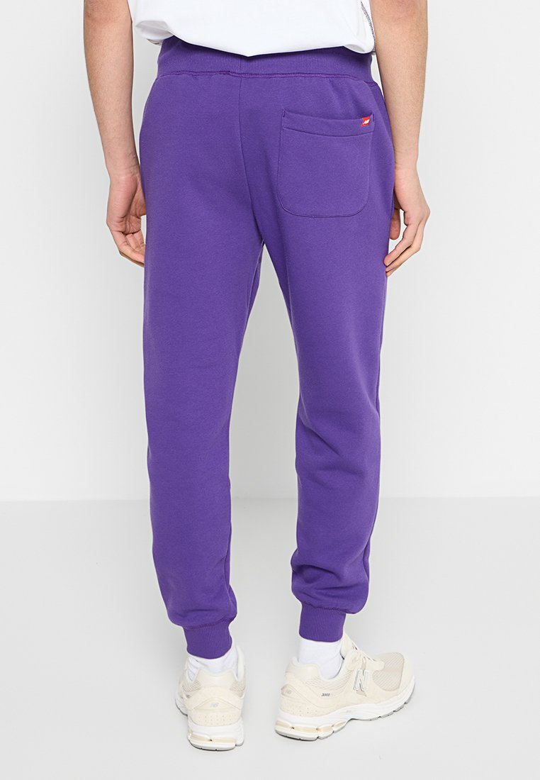 Persona che indossa pantaloni jogger viola con una tasca posteriore e scarpe da ginnastica bianche, in piedi davanti a uno sfondo bianco.