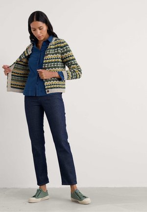 Frau trägt ein blaues Hemd mit Knöpfen, gemusterten Cardigan, dunkle Jeans und grüne Sneaker, hält den Cardigan offen und schaut nach unten.