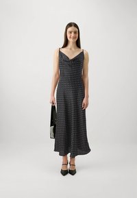 Γυναίκα που φοράει ένα μαύρο μιντι φόρεμα με λευκές βούλες, μαύρες γόβες και κρατά μια μαύρη και λευκή τσάντα, standing σε απλό λευκό φόντο.