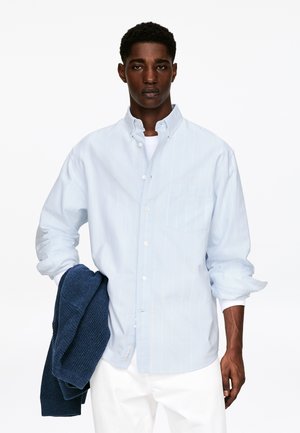 Ung mand iført en lyseblå skjorte med knapper over en hvid t-shirt, der holder en mørkeblå strikket sweater i venstre hånd mod en ensfarvet baggrund.