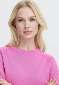 Femme blonde aux yeux bleus portant un pull rose vif, les bras croisés, regardant droit devant elle sur un fond clair.