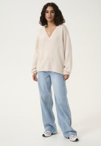 Pull en v couleur beige clair avec un tissu texturé, à manches longues, associé à un jean taille haute bleu clair et des baskets grises.