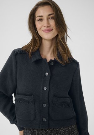 Schwarze strukturierte Jacke mit Kragen, Frontknopfverschluss und zwei mit Fransen versehenen Taschen. Besitzt ein dezentes Fischgrätmuster.