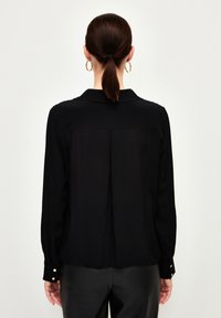 Camicia nera a maniche lunghe con colletto e dettagli in oro sui polsini. Tessuto leggero, texture liscia e dettaglio sul dorso.