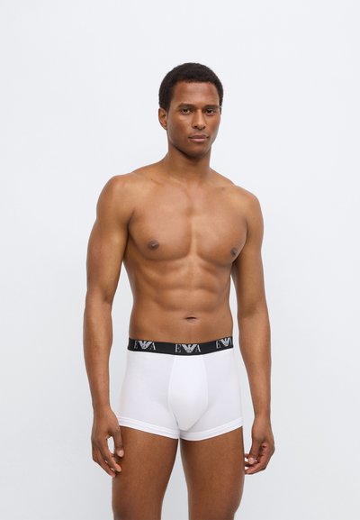 Λευκά μποξεράκια με μαύρη ελαστική μέση που φέρει λογότυπο. Το ύφασμα φαίνεται λείο, εφαρμοστό και ελαστικό. Το μοντέλο στέκεται.