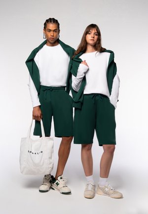 Grüne Baumwollshorts kombiniert mit weißen Langarmhemden, ergänzt durch einen passenden grünen Hoodie. Eine weiße Tragetasche mit schwarzem Text.