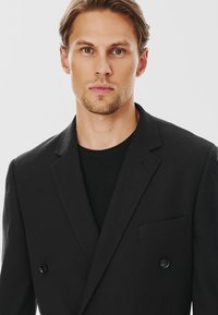 Czarny dwurzędowy żakiet z teksturowanej tkaniny, z klapami w szpic, pojedynczą kieszenią na piersi i dwoma guzikami z przodu. Noszony na czarnej koszuli.