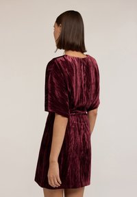 Kobieta z włosami do ramion w brązowym kolorze, nosząca krótką burgundową sukienkę z aksamitnego materiału i szerokimi rękawami, widziana z tyłu na jednolitym tle.