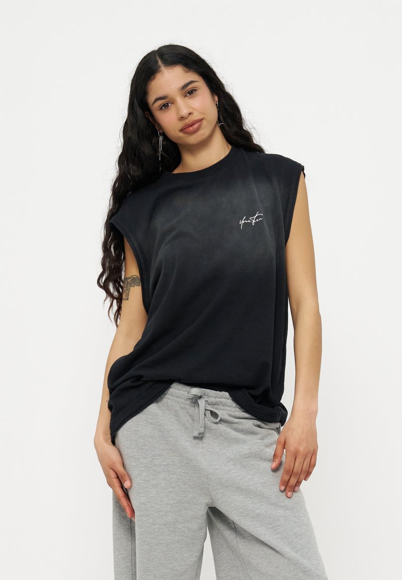 Γυναίκα με μακριά σγουρά μαλλιά, φοράει μαύρη αμάνικη μπλούζα με κείμενο και ανοιχτό γκρι παντελόνι φόρμας, ποζάροντας μπροστά σε απλό φόντο.