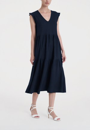 Donna che indossa un abito midi blu navy senza maniche con scollo a V e gonna a balze, abbinato a sandali bianchi con tacco alto e cinturini.