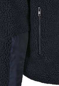 Mustast fliisist jakk, millel on tekstuurse, karvase välispinnaga ja sile satiinist varrukas nähtava tõmblukuga.