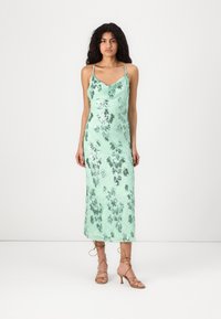 Mintgrünes, ärmelloses Satin-Maxikleid mit Blumenmuster und gewelltem Ausschnitt. Das Model trägt dazu leichte, braune Riemchenschuhe.