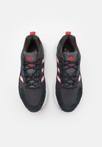 Svart- och gråa sportskor med röda och vita ränder, en texturerad ovandel och en gummisula. Kommer med svarta skosnören och en logotyp på tungan.