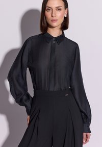 Czarna bluzka z długim rękawem, kołnierzykiem i guzikami. Gładka faktura, lekko prześwitujący materiał. W połączeniu z plisowanymi czarnymi spodniami.