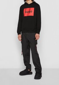 Felpa nera con cappuccio e logo rosso 'HUGO', abbinata a pantaloni cargo neri con tasche laterali e dettagli rossi, e sandali neri slide.
